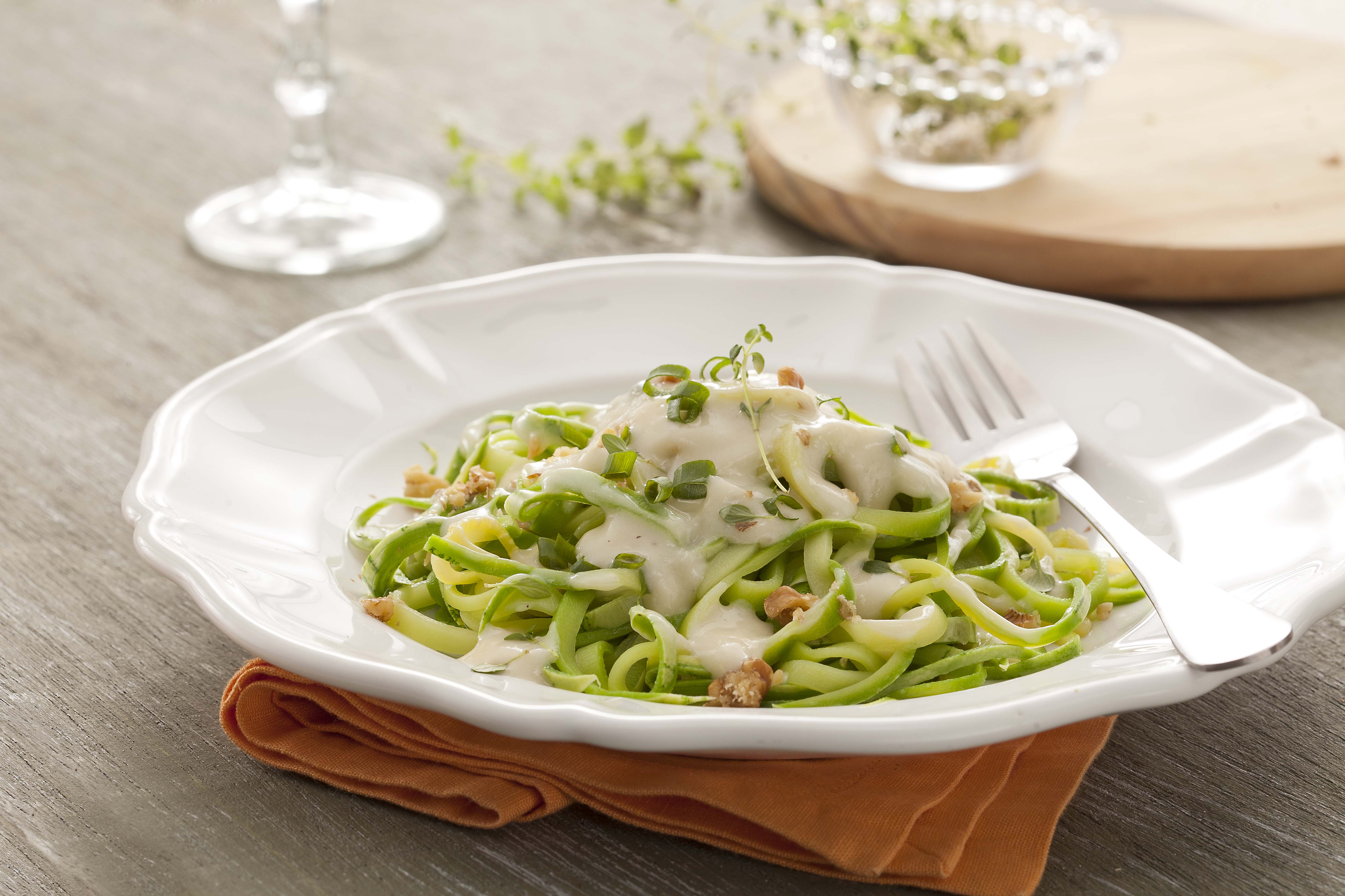 Fotografía en tonos verdes y blancos, en el centro un plato blanco profundo con espaguetis de calabacín verde en el interior con salsa blanca. Debajo del plato hay una servilleta de tela naranja sobre una mesa de madera.