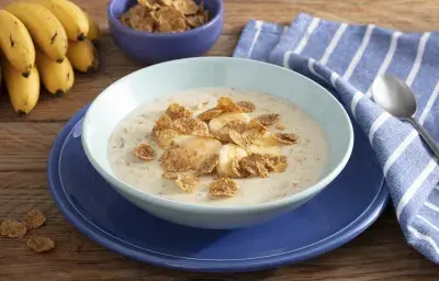 Fotografía en tonos de azul sobre un banco de madera con un paño azul con rayas blancas, un gran plato azul en el centro, con un plato hondo celeste y la papilla con cereales. Al fondo, un racimo de plátanos y una pequeña olla de cereal.