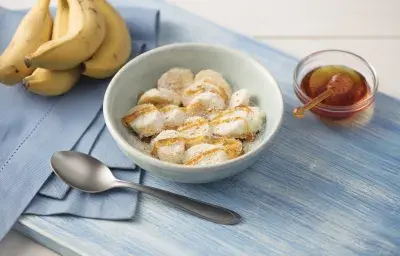 Fotografía en tonos de azul sobre una mesa de madera clara con un mantel azul al lado, un plato blanco profundo con rodajas de plátano cubiertas de miel, avena, leche en polvo y linaza. Junto a él, un racimo de plátanos y un botecito de miel.