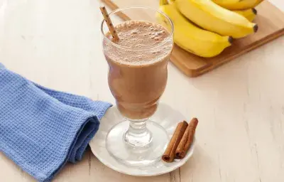 Fotografía en tonos marrones en un banco de madera clara con un paño azul al lado y en el centro un pequeño plato blanco con una taza de vidrio y el batido de cacao y plátano adentro. Al fondo, un racimo de plátanos apoyado en una tabla de madera.