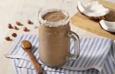 Paño rayado celeste y blanco con cuchara de madera marrón con mantequilla de maní. Una taza transparente con el batido y el borde de coco rallado.