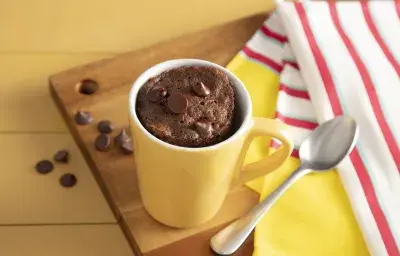 Fotografía en tonos amarillos sobre un banco de madera amarillo, una tela a rayas rojas y amarillas y una tabla de madera en el centro de la foto con una taza amarilla con el pastel de chocolate descansando sobre ella.