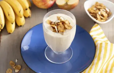 Imagen de un vaso con bebida cremosa en tono claro, decorado con cereal. Está en un plato azul, y alrededor hay cereales en la mesa, un racimo de plátanos, dos manzanas y una tela de rayas amarillas y blancas.