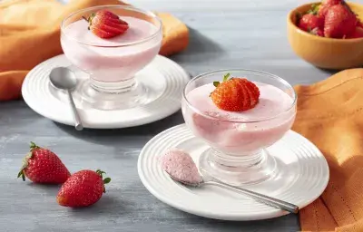 Imagen de primer plano de la receta de Strawberry Mousse Zero Lactose, en rosa claro, servida en frascos de vidrio, decorada con rodajas de fresas. La receta está en platos blancos sobre un mostrador con una tela naranja y más fresas enteras decorando