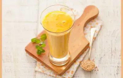 Fotografía en tonos amarillos y blancos tomada desde un banco visto de frente, contiene una tabla de madera y un vaso transparente con una bebida de mango encima.