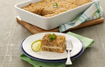 Fotografía en tonos de verde sobre un banco de madera gris, una tabla de cortar de madera, un paño de cocina verde, uno rayado en verde. En el centro, un plato redondo blanco con un trozo de kebab relleno. Al fondo, un refractario con toda la kibbeh.