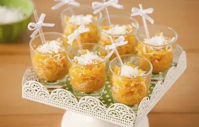 Fotografía en tonos naranjas sobre una mesa de madera con un puesto de caramelos blancos en el centro con caramelos de calabaza en vasos transparentes con palillos chinos y un lazo blanco.