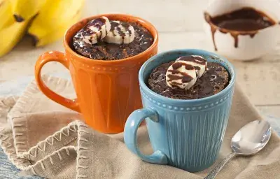 Fotografía en tonos beige, azul y naranja de una encimera blanca con un paño beige, sobre ella una taza azul y una taza naranja con pastel de taza. En el fondo un racimo de plátanos y un recipiente redondo con almíbar.