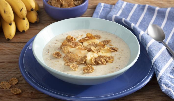 Fotografía en tonos de azul sobre un banco de madera con un paño azul con rayas blancas, un gran plato azul en el centro, con un plato hondo celeste y la papilla con cereales. Al fondo, un racimo de plátanos y una pequeña olla de cereal.