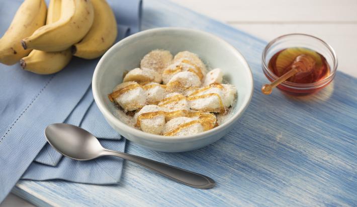 Fotografía en tonos de azul sobre una mesa de madera clara con un mantel azul al lado, un plato blanco profundo con rodajas de plátano cubiertas de miel, avena, leche en polvo y linaza. Junto a él, un racimo de plátanos y un botecito de miel.