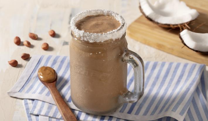 Paño rayado celeste y blanco con cuchara de madera marrón con mantequilla de maní. Una taza transparente con el batido y el borde de coco rallado.