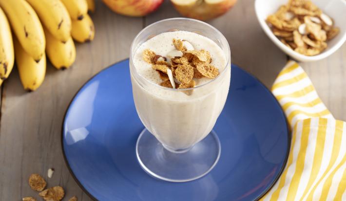 Imagen de un vaso con bebida cremosa en tono claro, decorado con cereal. Está en un plato azul, y alrededor hay cereales en la mesa, un racimo de plátanos, dos manzanas y una tela de rayas amarillas y blancas.