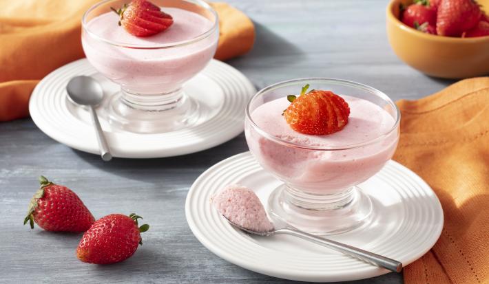 Imagen de primer plano de la receta de Strawberry Mousse Zero Lactose, en rosa claro, servida en frascos de vidrio, decorada con rodajas de fresas. La receta está en platos blancos sobre un mostrador con una tela naranja y más fresas enteras decorando