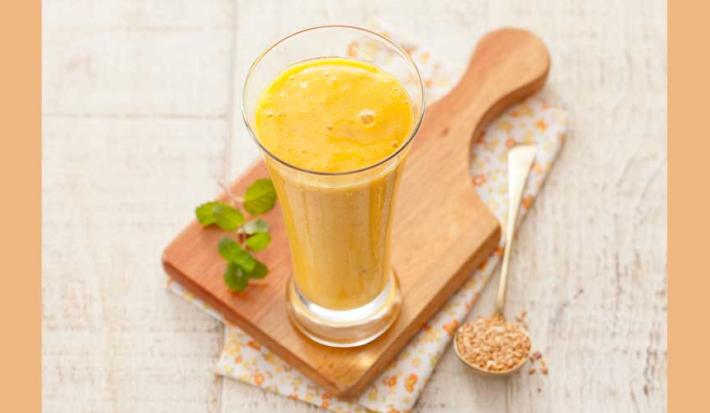 Fotografía en tonos amarillos y blancos tomada desde un banco visto de frente, contiene una tabla de madera y un vaso transparente con una bebida de mango encima.