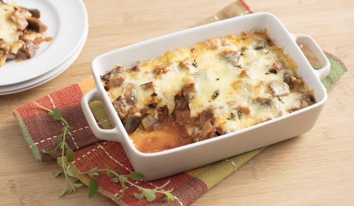 Fotografía en tonos blancos y amarillos de encimeras de madera clara, con tela a cuadros rojos y verdes, fuente rectangular blanca con asas y trozos de berenjena y queso en su interior. Al fondo, tres platos blancos apilados con un trozo de la receta.