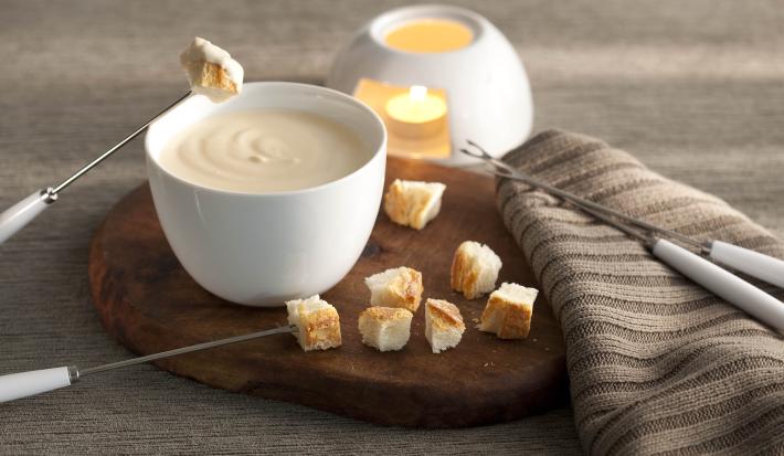 fotografía en tonos grises, marrones y banco visto de frente. Contiene una tabla de madera redonda con un recipiente blanco redondo que contiene la fondue, 4 tenedores de fondue y trozos de pan alrededor.