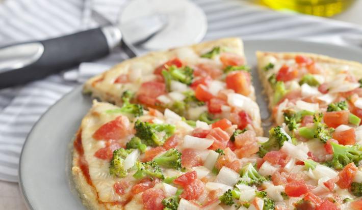 La fotografía en tonos blancos de una encimera de vista frontal contiene un plato blanco redondo con una pizza con trozos de tomate y brócoli en el fondo y una jarra de aceite.