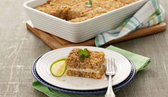 Fotografía en tonos de verde sobre un banco de madera gris, una tabla de cortar de madera, un paño de cocina verde, uno rayado en verde. En el centro, un plato redondo blanco con un trozo de kebab relleno. Al fondo, un refractario con toda la kibbeh.