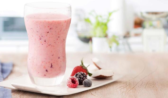 fotografía y tonos de rosa y blanco de un banco beige visto de frente, contiene un vaso transparente con la bebida de frutos rojos y al lado contiene frutos del bosque.