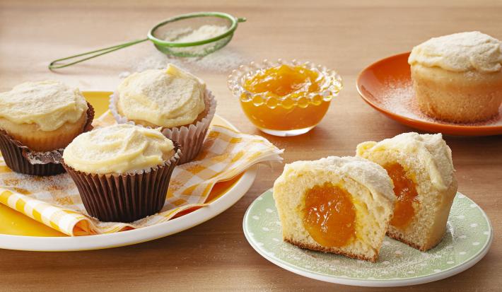 Fotografia em tons de laranja em uma bancada de madeira com um prato branco com paninho listrado em amarelo e três cupcakes de leite Ninho com manga em cima dele. Ao lado, um prato branco com um cupcake cortado ao meio mostrando o recheio de manga.